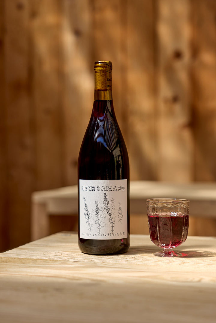 A bottle of Broc Cellars 2024 Negroamaro with a botanical label sits on a light wooden table beside a glass of red wine. Blurred warm wood paneling forms the background. This exquisite vintage is coming soon, promising rich flavor and elegance.