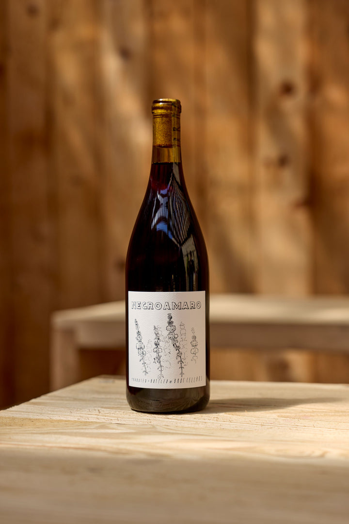 A bottle of Broc Cellars 2024 Negroamaro rests on a light wooden table with a wooden background. Its label features a simple botanical illustration. Coming soon.