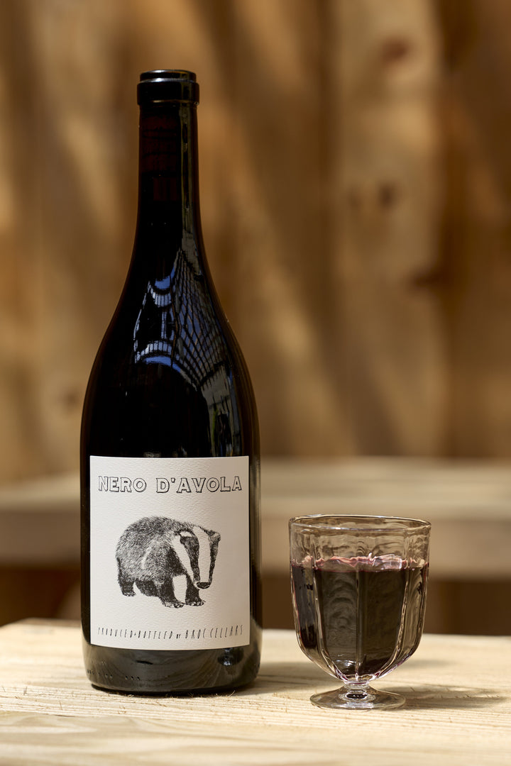 A bottle of Broc Cellars 2023 Nero D’Avola with a badger illustration on the label next to a small glass of red wine.