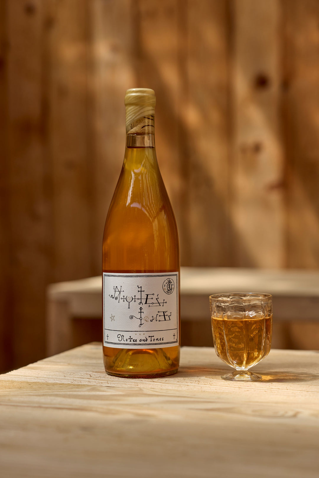 A bottle of 2022 Notes &amp; Tones - The Wine by Broc Cellars, with a handwritten-style label, sits on a light wooden table near a faceted glass. Sunlit wooden panels provide a warm backdrop for this naturally fermented wine.