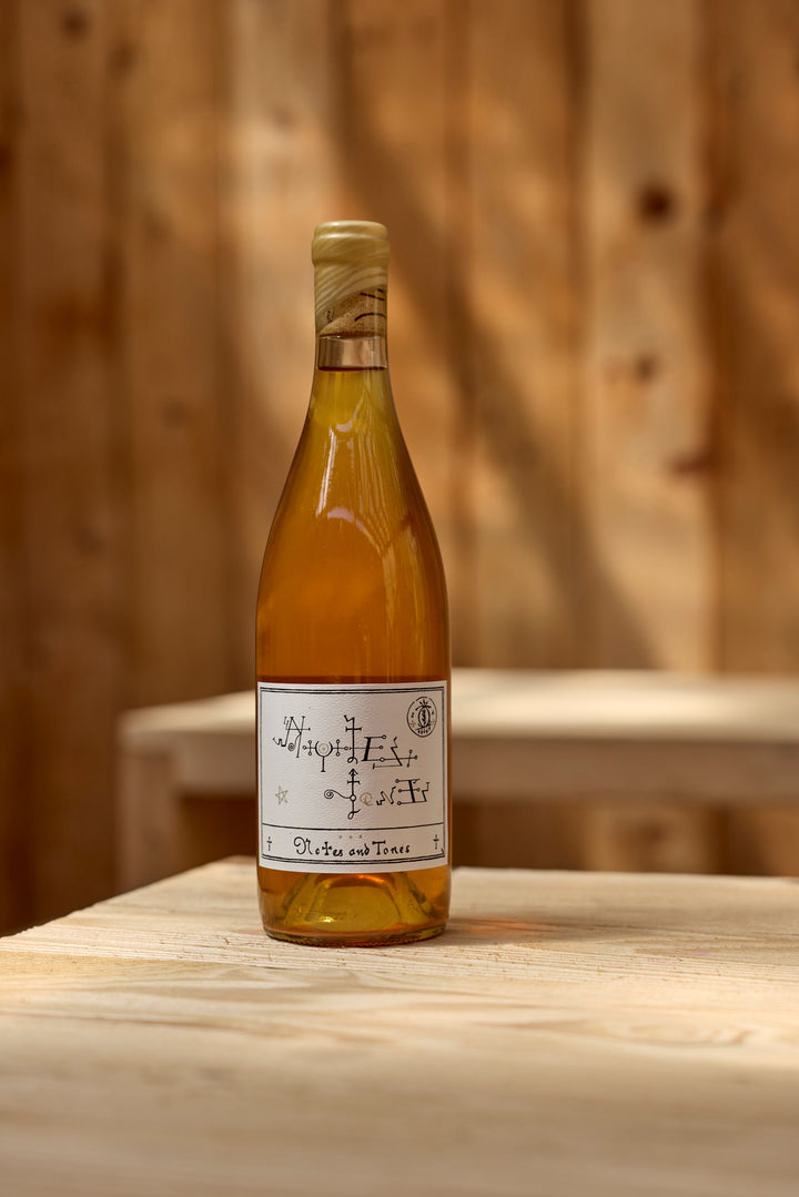 A bottle of Broc Cellars 2022 Notes &amp; Tones - The Wine, featuring a handwritten-style white label, rests on a light wooden table with a blurred wooden wall in the background. This wine is naturally fermented from organic grapes.