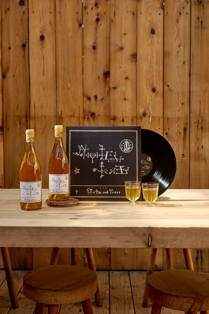 Two bottles and two glasses of Broc Cellars’ Notes &amp; Tones V.2—The Album and Wine Set sit on a wooden table beside a black vinyl record and its cover, creating a warm, rustic scene against a wooden wall backdrop.