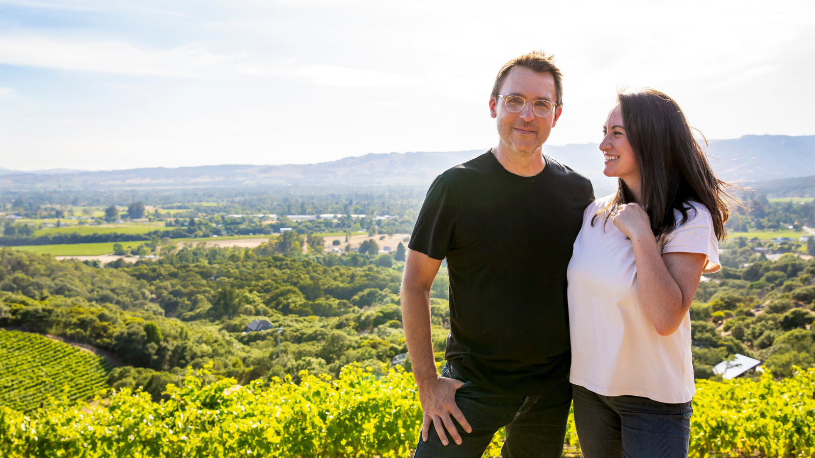 A man and woman stand before Fox Hill Vineyard’s rolling hills. He wears glasses and black; she smiles in white. They enjoy The Fox Hill Collection from Broc Cellars, crafted from estate-grown Italian varietals on this organic property.