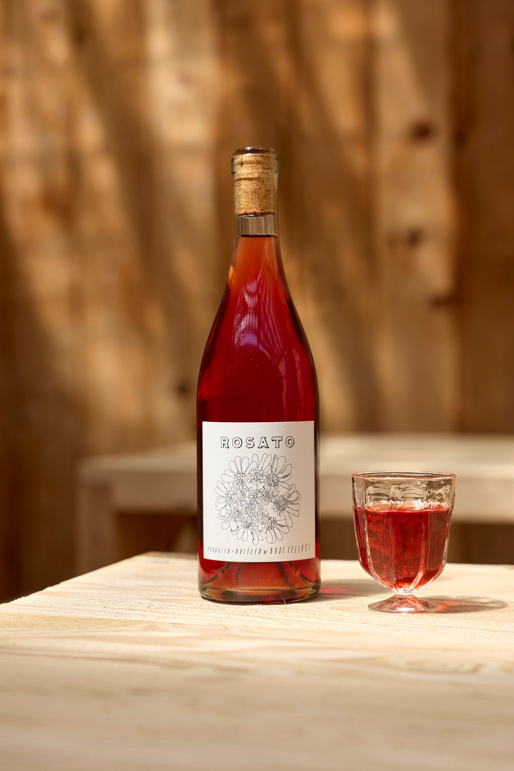 A bottle of Broc Cellars 2025 Rosato with a floral label sits on a light wooden table beside a glass of rosé wine and a tech sheet, set against softly blurred wood paneling in the background.
