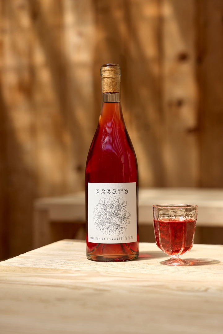 A bottle of Broc Cellars 2025 Rosato with a floral label sits on a light wooden table beside a glass of rosé wine and a tech sheet, set against softly blurred wood paneling in the background.