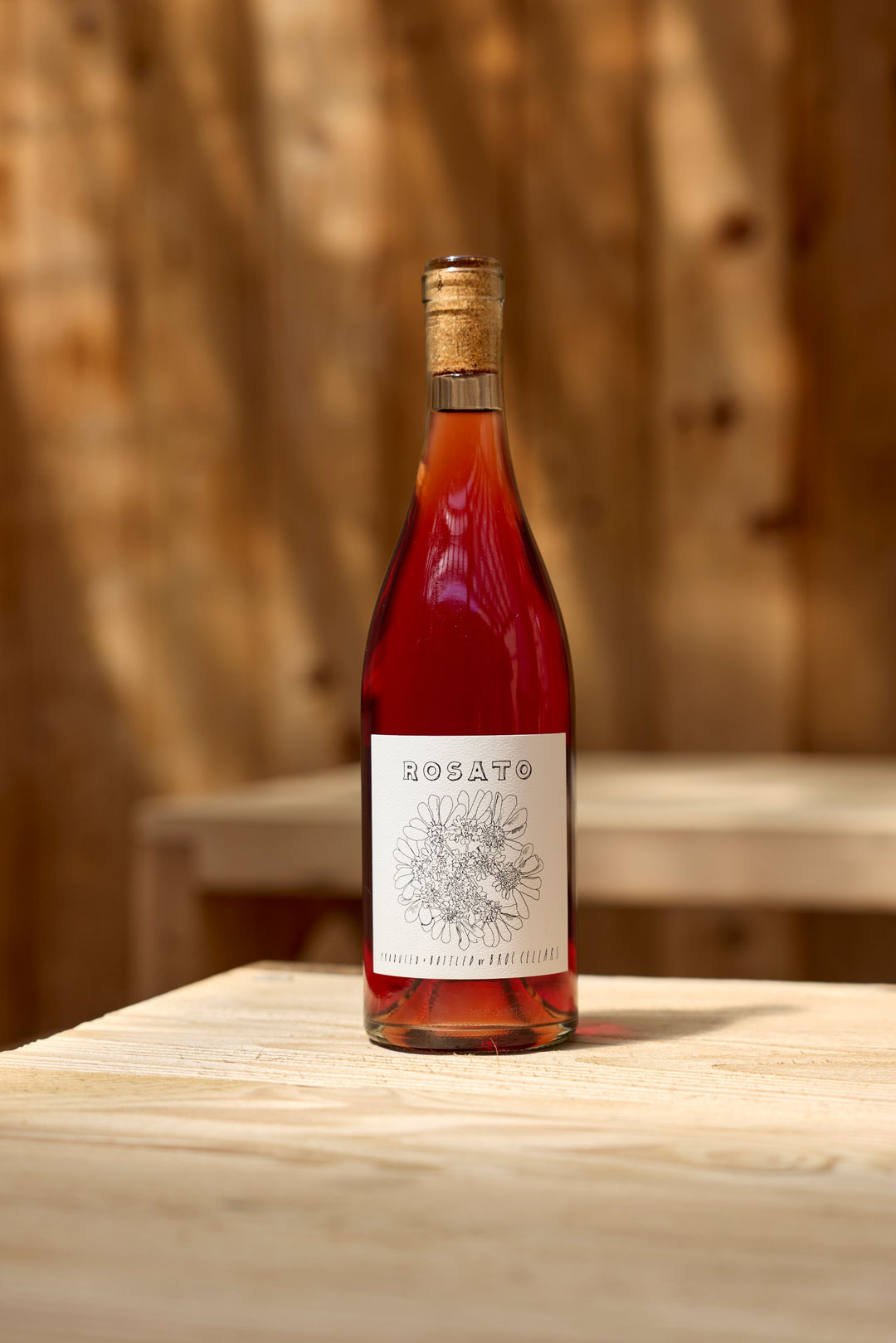 A bottle of 2025 Rosato by Broc Cellars, with a black and white floral label, sits on a light wooden table against a blurred wooden backdrop—ideal for your next tasting or exploring its tech sheet.