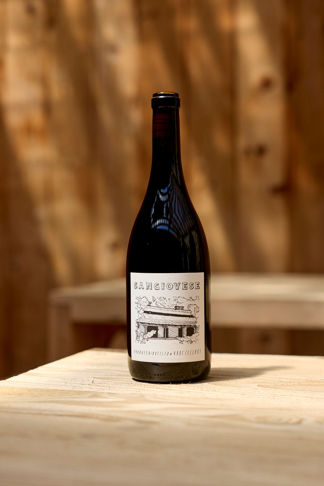 A bottle of 2025 Sangiovese from Broc Cellars rests on a light wooden table against a softly blurred wooden backdrop. The label showcases a black and white illustration of a building along with the name “Sangiovese.” Renowned for its food-friendly acidity and gentle tannins, this California wine is an excellent companion to a wide range of dishes.