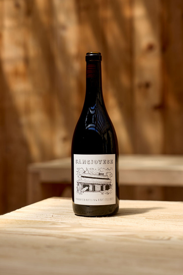 A bottle of 2025 Sangiovese from Broc Cellars rests on a light wooden table against a softly blurred wooden backdrop. The label showcases a black and white illustration of a building along with the name “Sangiovese.” Renowned for its food-friendly acidity and gentle tannins, this California wine is an excellent companion to a wide range of dishes.