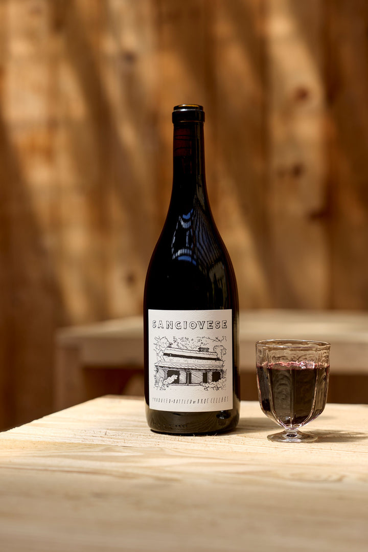 A bottle of 2023 Sangiovese from Broc Cellars and a small glass of California wine rest on a light wooden table, set against a blurred wood background. The label shows an illustration of a building.