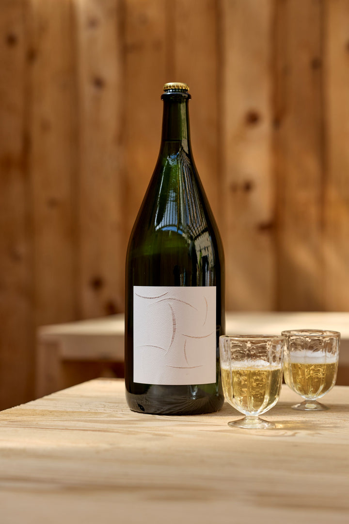 A large green bottle of Broc Cellars 2024 Sparkling Chenin Magnum with a minimalist white label sits on a light wooden table beside two glasses of pale sparkling wine, against a blurred wooden wall background.