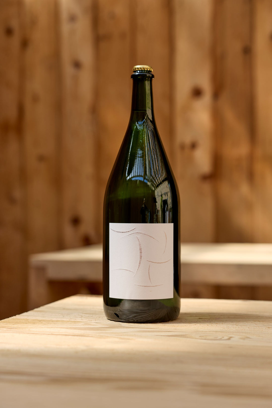 A large green glass bottle of Broc Cellars 2024 Sparkling Chenin Magnum with a blank white label sits on a light wooden table, set against a wooden wall background.