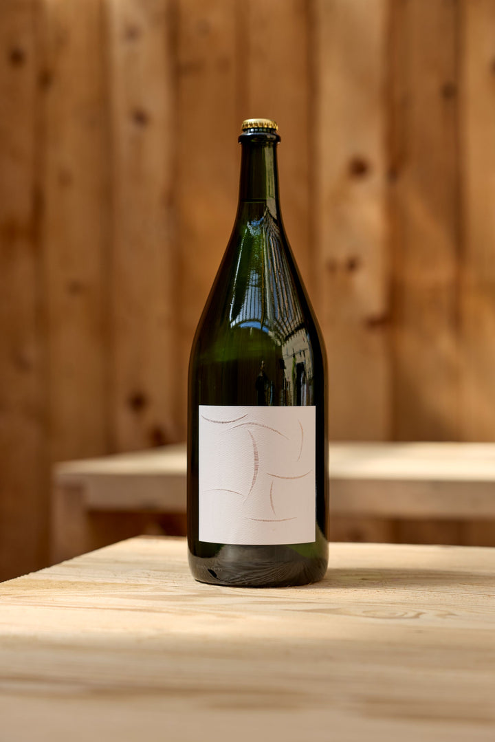 A large green glass bottle of Broc Cellars 2024 Sparkling Chenin Magnum with a blank white label sits on a light wooden table, set against a wooden wall background.