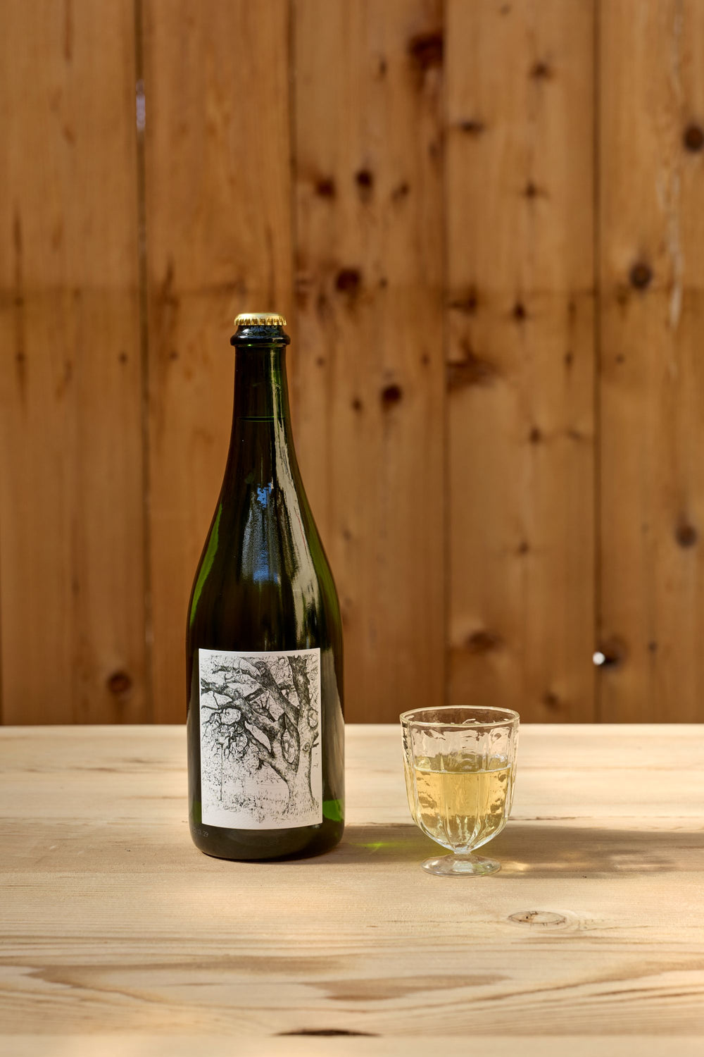 A green bottle of Broc Cellars 2025 Sparkling Erbaluce with a black and white illustrated label stands on a wooden table next to a small glass filled with a light golden beverage. The background is an unpainted wooden wall, creating the perfect rustic setting for this hand-picked selection.