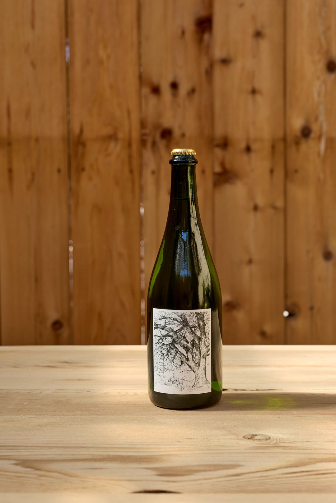 A green glass bottle of Broc Cellars 2025 Sparkling Erbaluce, featuring a black and white illustrated label, stands on a light wooden table in front of a wooden fence.