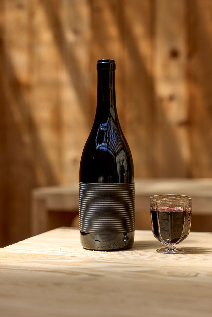 A bottle of 2023 Broc Valdiguié by Broc Cellars stands beside a filled glass on a light wooden table, set against a blurred wooden backdrop with soft, natural lighting.