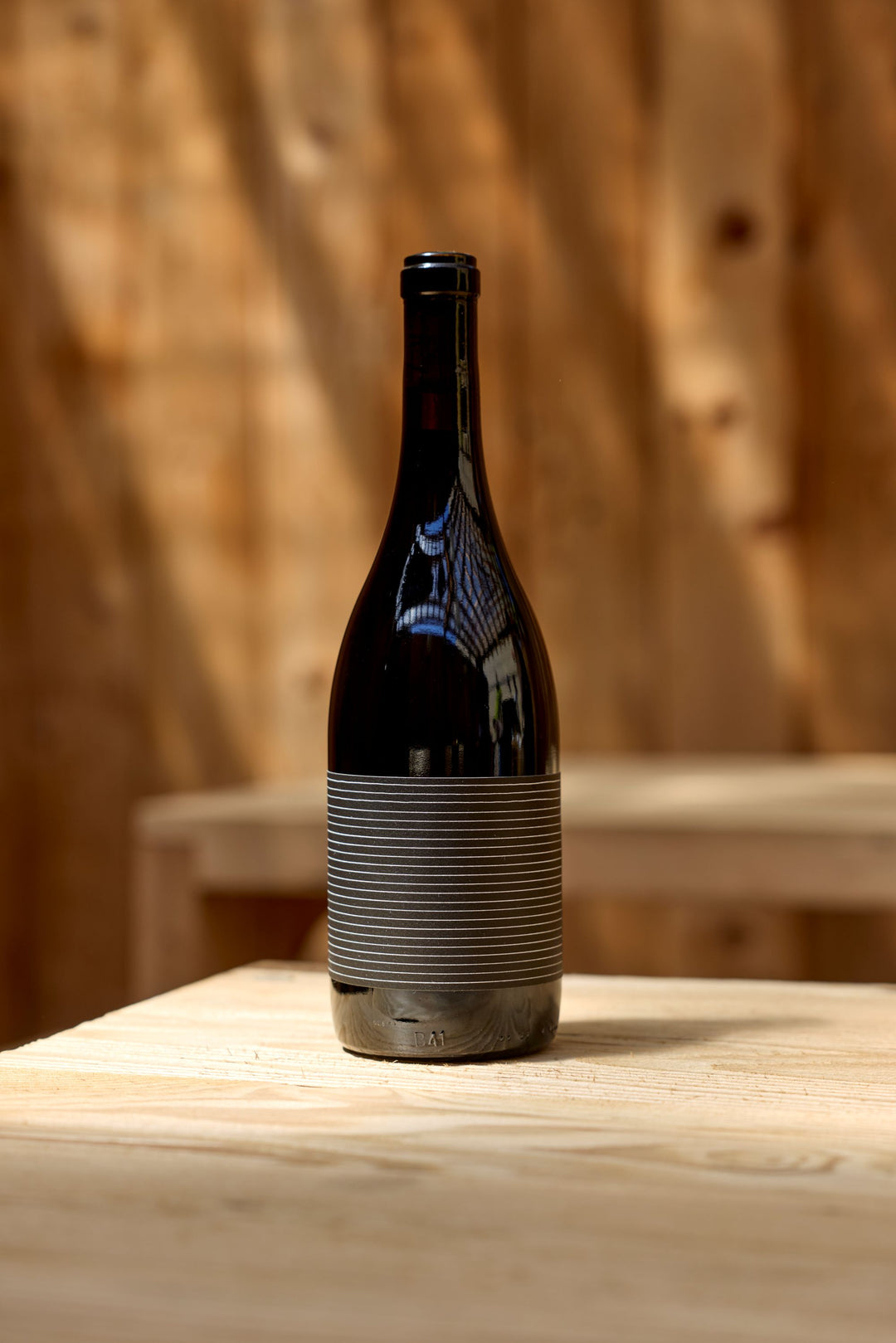 A dark glass bottle labeled with gray stripes sits on a light wood table, sunlight casting shadows behind it and highlighting the 2023 Broc Valdiguié from Broc Cellars.
