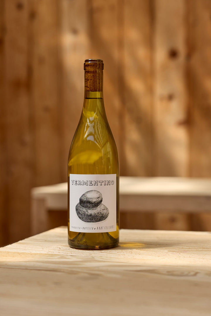 A bottle of 2023 Fox Hill Vermentino by Broc Cellars rests on a light wooden table with a blurred wooden wall behind it. The label showcases a black and white stone illustration with text.