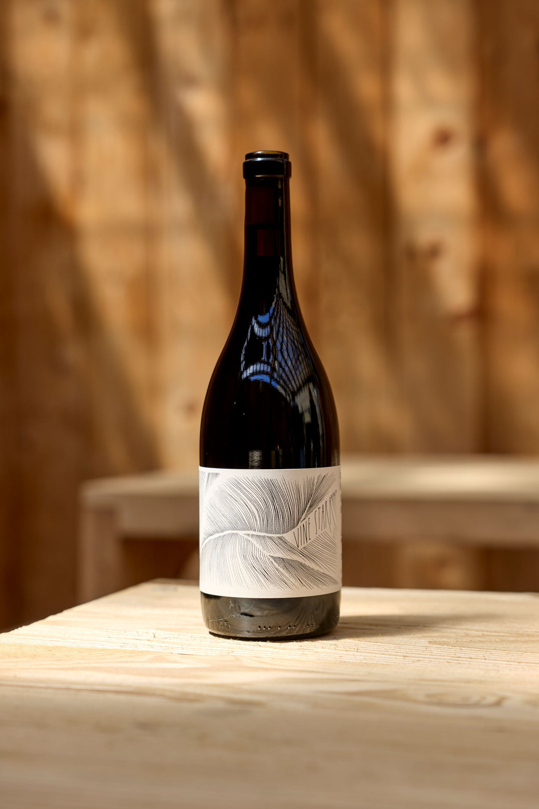 A dark glass wine bottle with a white label featuring a feather design sits on a light wooden table—a refined 2023 Vine Starr Zinfandel from Broc Cellars, set against a blurred wooden wall in the background.