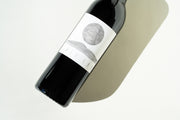 A wine bottle, 2023 Cali Cab by Broc Cellars, lies on its side on a light surface. The labels minimalist design with a stylized circle above a curved line hints at its California Cabernet Sauvignon origin. Shadows contrast against the background, promising rich wine from organically grown grapes.