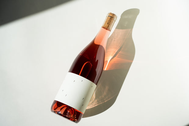 The 2024 Love Rosé from Broc Cellars, with a minimalistic white label, lies on its side casting a shadow. Sunlight accentuates the pink hue of the corked bottle, reminiscent of a delicate Valdiguié.