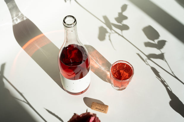 A bottle of 2024 Love Rosé by Broc Cellars and a filled glass rest on a white surface. A cork is nearby, while leaf and stem shadows, cast by sunlight from the left, create an inviting ambience.