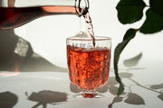 A pinkish-red 2024 Love Rosé from Broc Cellars is poured into a textured glass, forming bubbles. In the background, a silhouette of leaves and a rose stem can be seen.