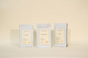 Three white bags labeled “Genmaicha,” “Mountain Beauty,” and “Sencha” from Broc Cellars’ Mothers Day Packs stand side by side against a neutral background, each featuring minimalist labels with additional text beneath the names.