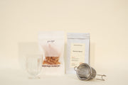 The Broc Cellars Mothers Day Packs feature a clear glass, dried orange candied peel, Mountain Beauty Black Tea in a sealed packet, and a metal infuser arranged on a light beige surface.