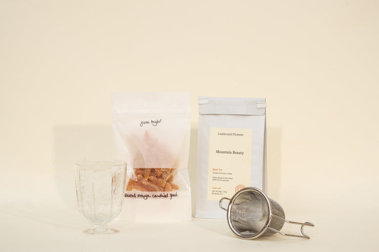 The Broc Cellars Mothers Day Packs feature a clear glass, dried orange candied peel, Mountain Beauty Black Tea in a sealed packet, and a metal infuser arranged on a light beige surface.