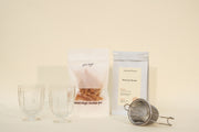 The Broc Cellars Mothers Day Packs include two clear glass cups, Mountain Beauty black tea, dried orange candied peel, and a metal tea strainer arranged on a light surface with a plain background.