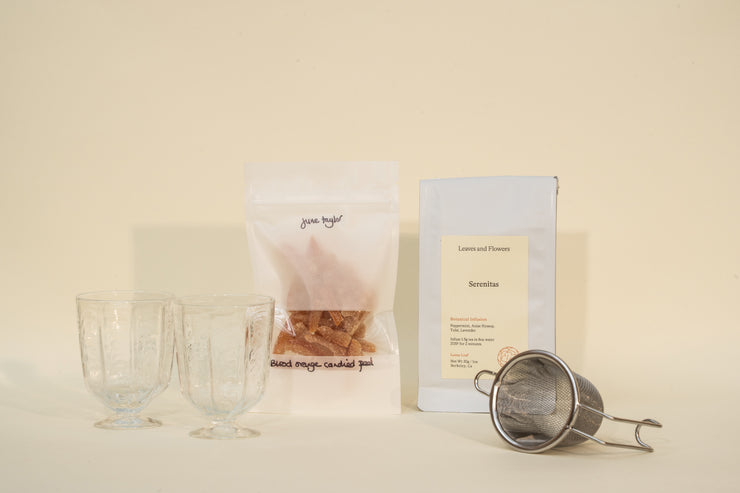 The Mothers Day Packs by Broc Cellars feature two clear glass cups, dried mango pieces, a white Serenitas tea bag, and a metal tea infuser displayed on a light surface against a pale background.