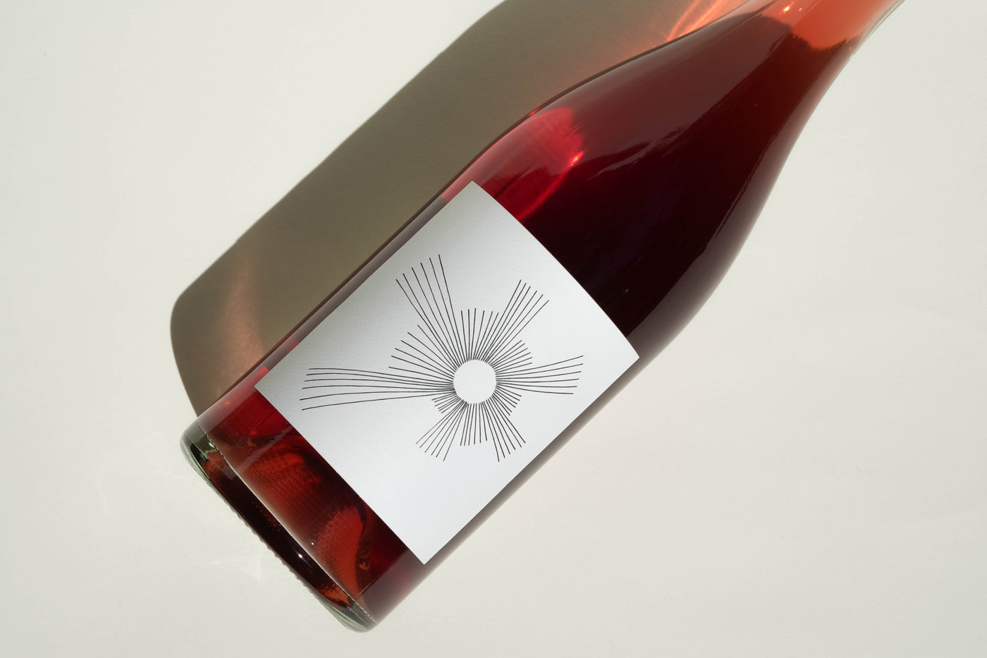 A glass bottle of 2024 Oh Sunshine rosé by Broc Cellars, featuring a modern white label with minimalist black lines radiating from a central circle, rests on a light surface casting a shadow.