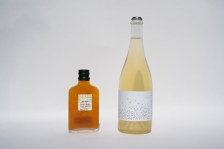 A small amber Clementine + Lemon Thyme Syrup bottle with a yellow label sits beside a tall clear Broc Cellars Spritz Pack bottle. The latter resembles Sparkling Chenin Blanc, with its white label and silver cap enclosing light yellow liquid, both set elegantly against a plain white background.