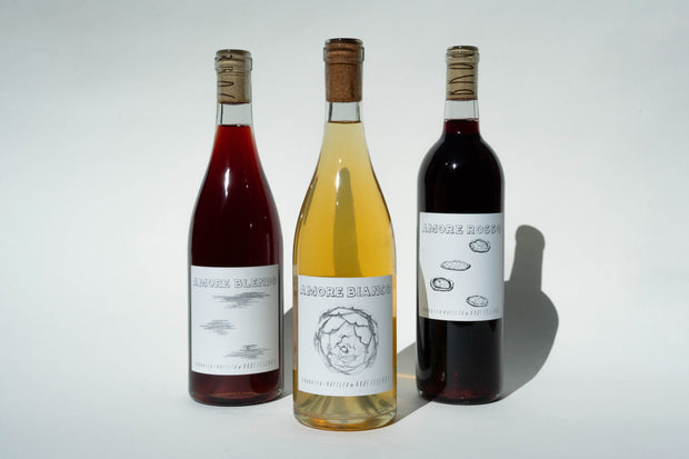 The Amore Trio from Broc Cellars includes three corked wine bottles with unique illustrated labels. This premium Italian set features distinct hues: a red, a light amber, and a deep red. The plain white background highlights the essence of these elegant table wines.