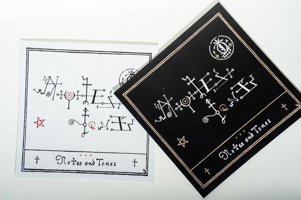 Two square items, part of the Broc Cellars Notes & Tones V.1 + V.2 - Album Duo, are displayed on a white surface. One is white with black and red abstract symbols, the other is black with similar designs, gold accents, and borders.