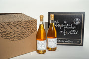 Two bottles of amber liquid with artistic labels sit beside a decorative black sign and a cardboard box with curved line patterns, echoing vintage vinyl aesthetics. Both feature abstract symbols and text reflecting Broc Cellars style. Product: Notes & Tones V.2 - The Album and Wine Set by Broc Cellars.