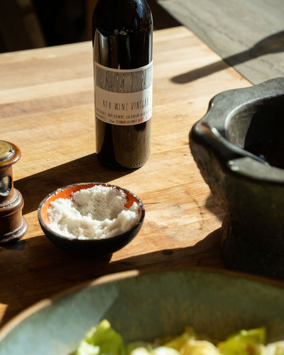 A bottle of red wine vinegar stands on a wooden surface next to a small bowl of salt, a pepper grinder, and a stone mortar. Part of a leafy salad is visible in the foreground. Sunlight casts shadows across the scene.