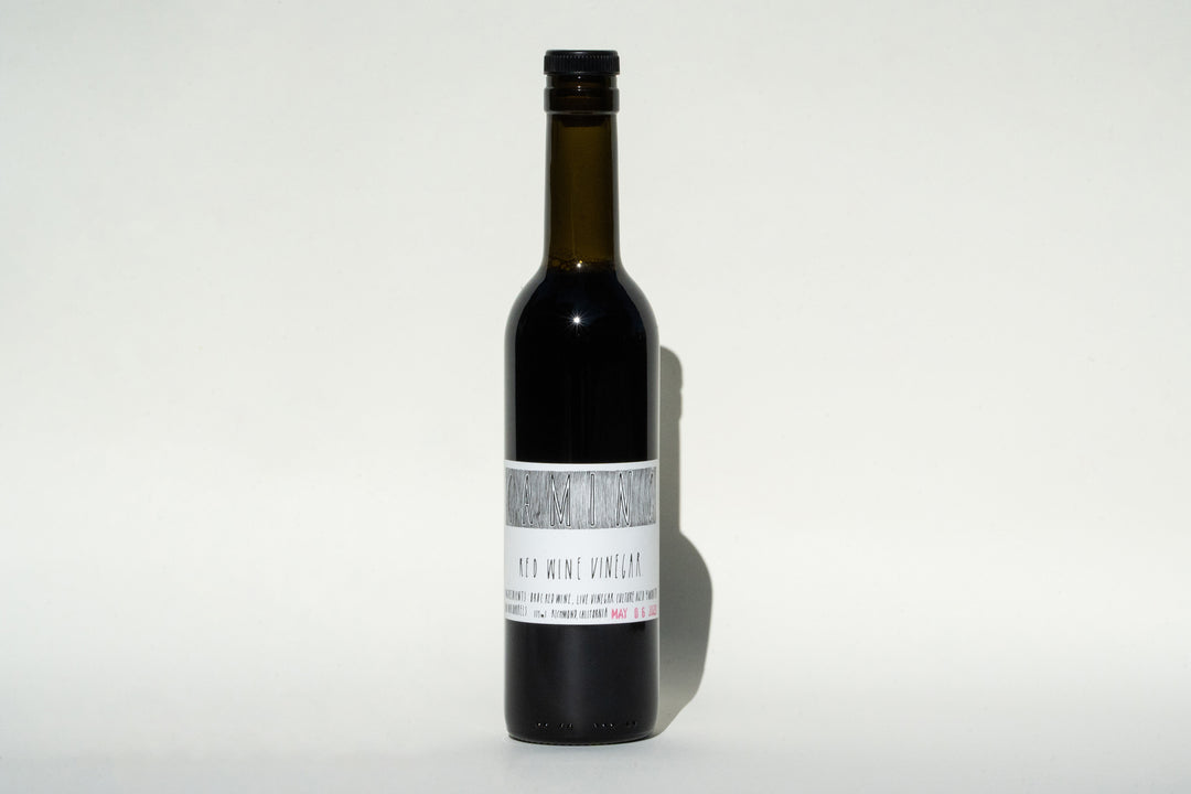 A dark glass bottle with a black cap stands against a plain background. It features a white label that reads Camino Red Wine Vinegar from Broc Cellars, suggesting organic farming practices. The bottle contains a deep-colored liquid, presumably the vinegar.