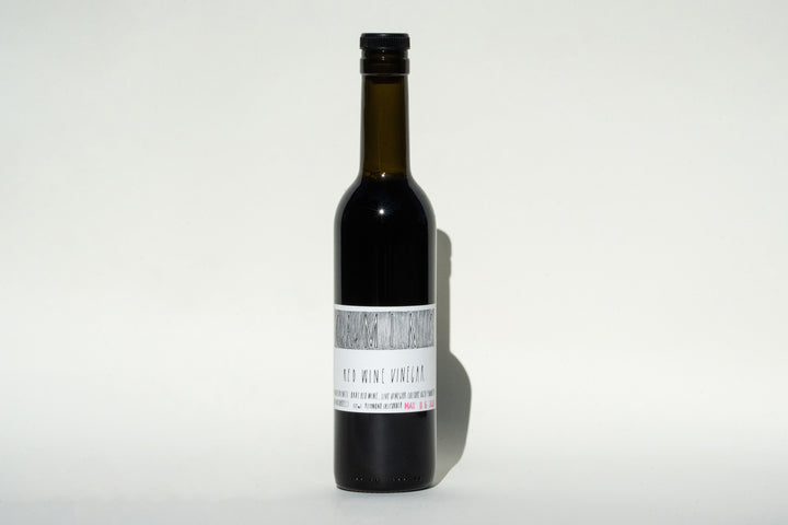 A dark glass bottle with a black cap stands against a plain background. It features a white label that reads Camino Red Wine Vinegar from Broc Cellars, suggesting organic farming practices. The bottle contains a deep-colored liquid, presumably the vinegar.