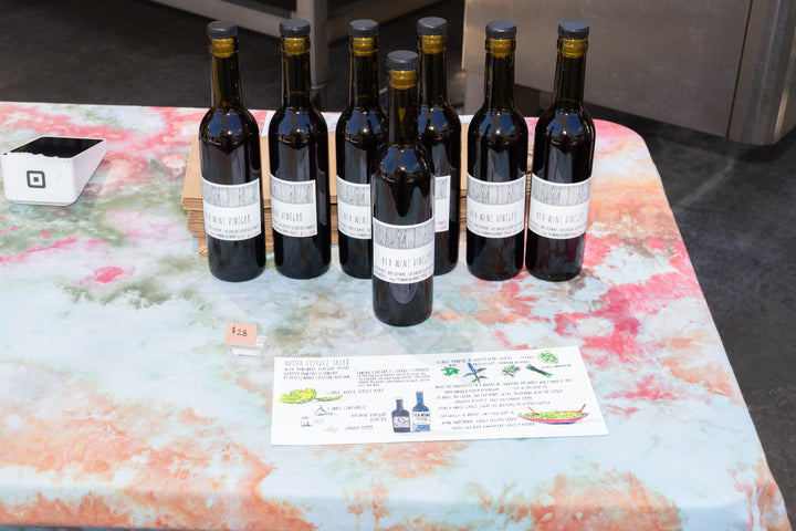 Eight dark glass bottles with white labels—CAMINO RED WINE VINEGAR by Broc Cellars—are displayed on a tie-dye tablecloth beside a $25 price tag and informational sheet. A white payment terminal is in the top left corner.