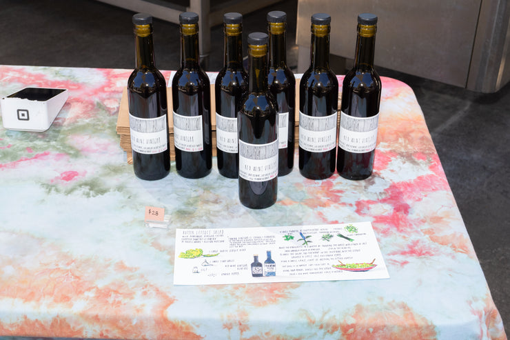 Eight dark glass bottles with white labels—CAMINO RED WINE VINEGAR by Broc Cellars—are displayed on a tie-dye tablecloth beside a $25 price tag and informational sheet. A white payment terminal is in the top left corner.