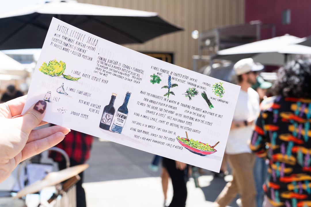 A person holds a colorful menu or recipe card outdoors at a lively market, with people, umbrellas, and food stands in the background—featuring CAMINO RED WINE VINEGAR from Broc Cellars.