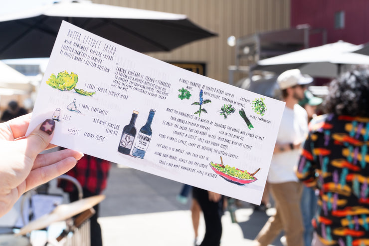 A person holds a colorful menu or recipe card outdoors at a lively market, with people, umbrellas, and food stands in the background—featuring CAMINO RED WINE VINEGAR from Broc Cellars.