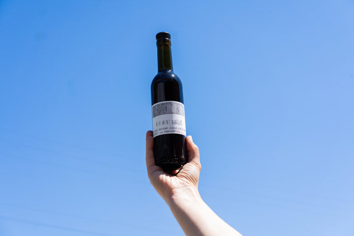 A hand holds up a dark glass bottle of Broc Cellars CAMINO RED WINE VINEGAR with a white label against a clear blue sky. The label is partly obscured and not fully readable.