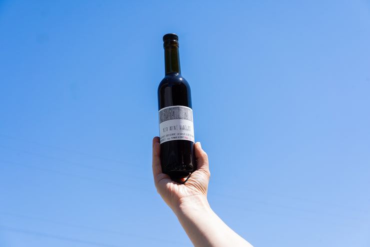 A hand holds up a dark glass bottle of Broc Cellars CAMINO RED WINE VINEGAR with a white label against a clear blue sky. The label is partly obscured and not fully readable.