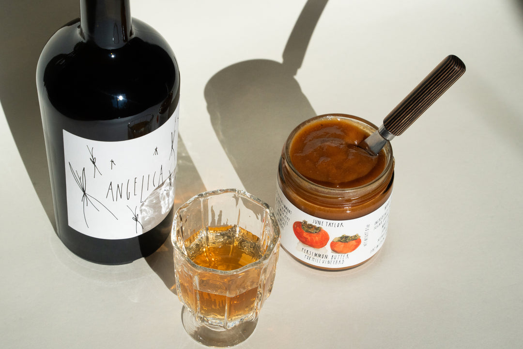 A bottle with a handwritten label, a glass filled with Broc Cellars golden liquid, and a jar labeled June Taylor Persimmon Butter containing a spoon are arranged on a white surface, casting elegant shadows.