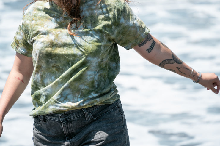 Outdoors, someone in a Broc Cellars UNA Ice Dyed Organic Short Sleeve Shirt | Coast and gray shorts displays several tattoos, including a snake. A blurred wavy body of water adds tranquility to the scene.