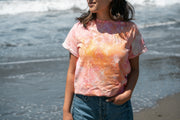 A person stands on a beach, partially facing the camera, wearing the Broc Cellars UNA Ice Dyed Organic Short Sleeve Shirt in Island colors and blue jeans. The ocean waves provide a serene backdrop. With shoulder-length hair and one hand in their pocket, they exude a relaxed vibe.