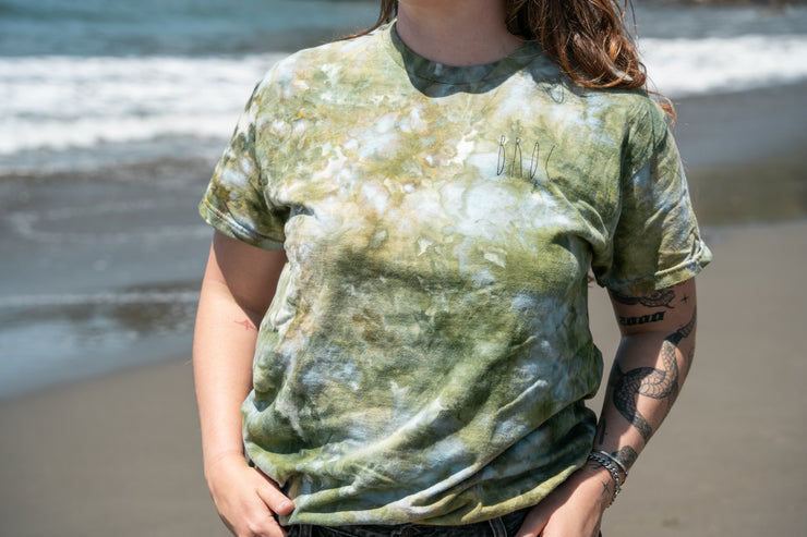 On the beach, a person sports a Broc Cellars UNA Ice Dyed Organic Short Sleeve Shirt in Coast and jeans, tattoos visible on their arms as they stand with hands in pockets. Gentle waves approach the sandy shoreline in the background.