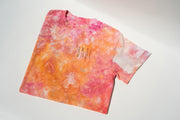 The UNA Ice Dyed Organic Short Sleeve Shirt by Broc Cellars, featuring pink, orange, and white shades with BRB in black on the front, is laid flat on a light surface. Made in the USA, soft shadows form around it.